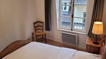 Une chambre à coucher avec un lit, une chaise en bois, une fenêtre avec des rideaux et un radiateur.
