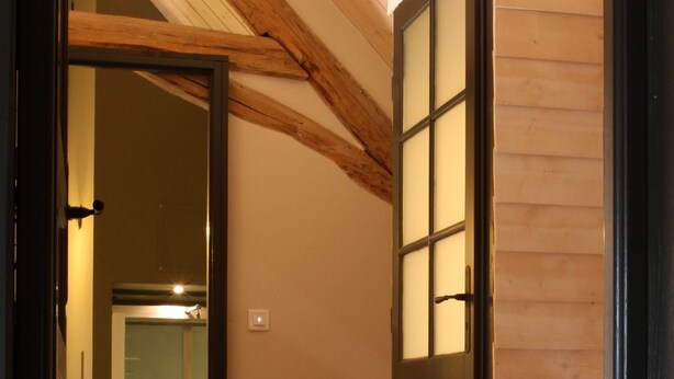 Vue intérieure d’une pièce avec des poutres en bois au plafond, une porte dotée de panneaux en verre dépoli et un plancher en bois.