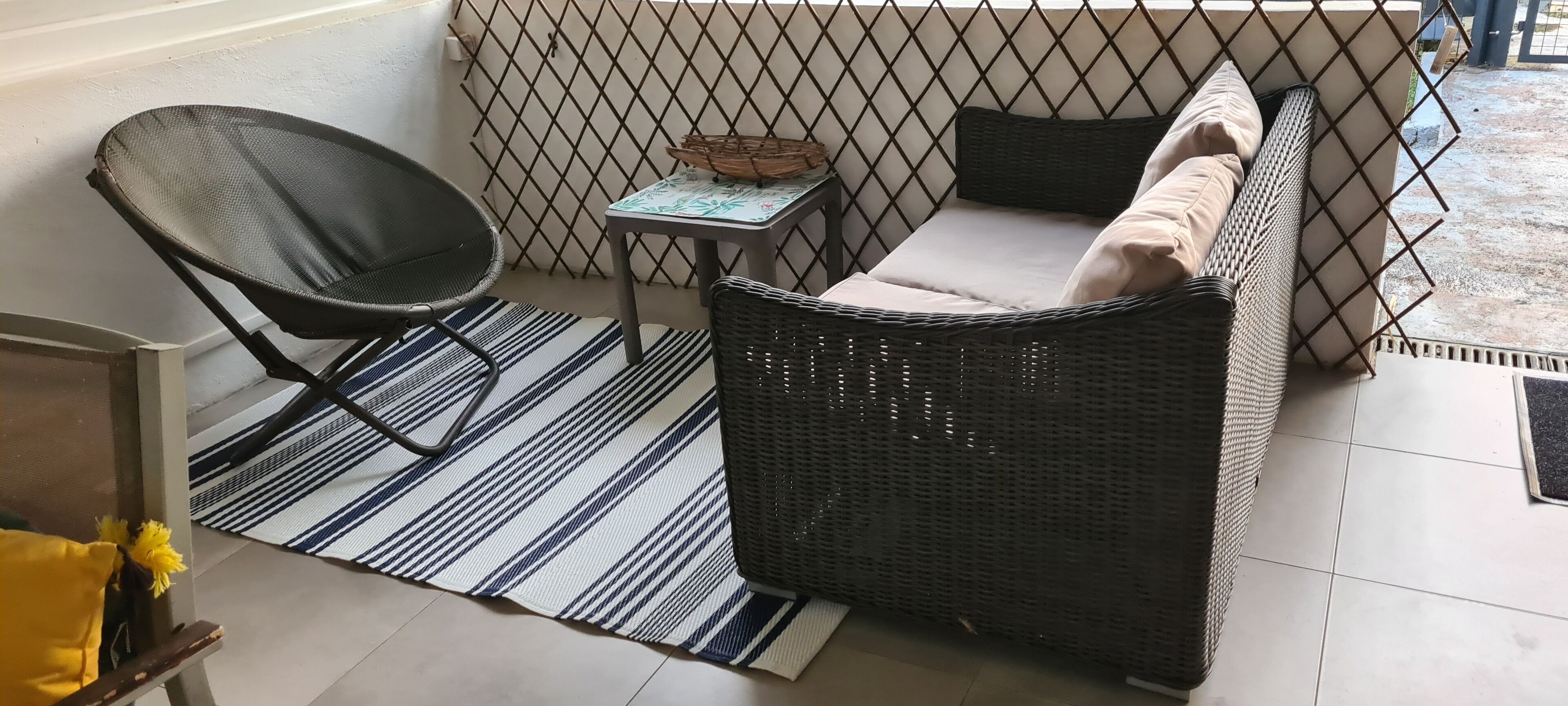 Ein Balkon mit einer Rattan-Couch, einem Sessel, einem kleinen Tisch und einer Streifenoptik-Isomatte.