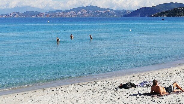 A beach with clear blue water, people swimming, and mountains in the background.