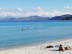 Ein Strand mit klarem, blauem Wasser, darin schwimmende Personen und Berge im Hintergrund.