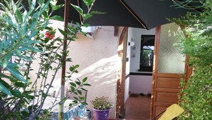 A covered outdoor seating area with a yellow chair, potted plants, and a wooden door leading to an interior space.