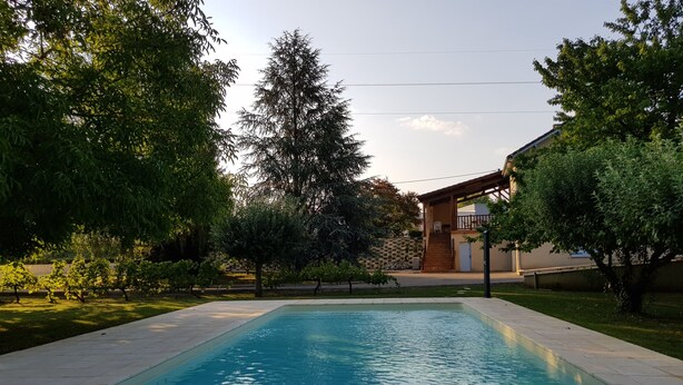 A rectangular swimming pool with a wooden deck in the foreground, surrounded by lush greenery and a building with a covered patio in the background. - Charming holiday home with private heated pool (Prayssac)