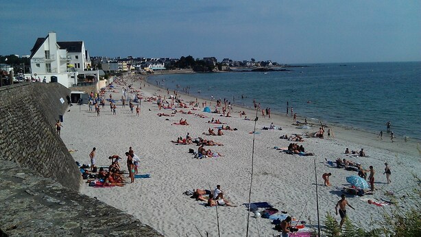 Une plage où des gens se prélassent au soleil et se baignent. On aperçoit un hôtel à l’arrière-plan, avec une enseigne visible.