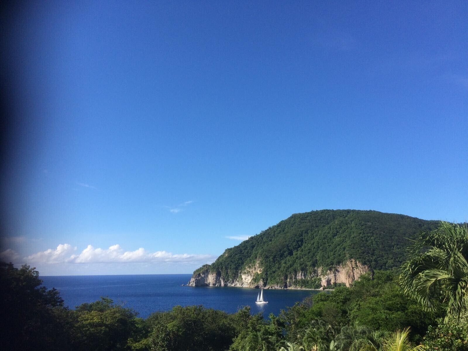 A coastal landscape with a clear view of the sea, a sailboat, and a lush green hill.