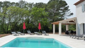 A rectangular swimming pool with lounge chairs and red umbrellas, surrounded by trees and a building with a tiled roof. - Air-conditioned villa with heated pool for 9 people (La Motte)
