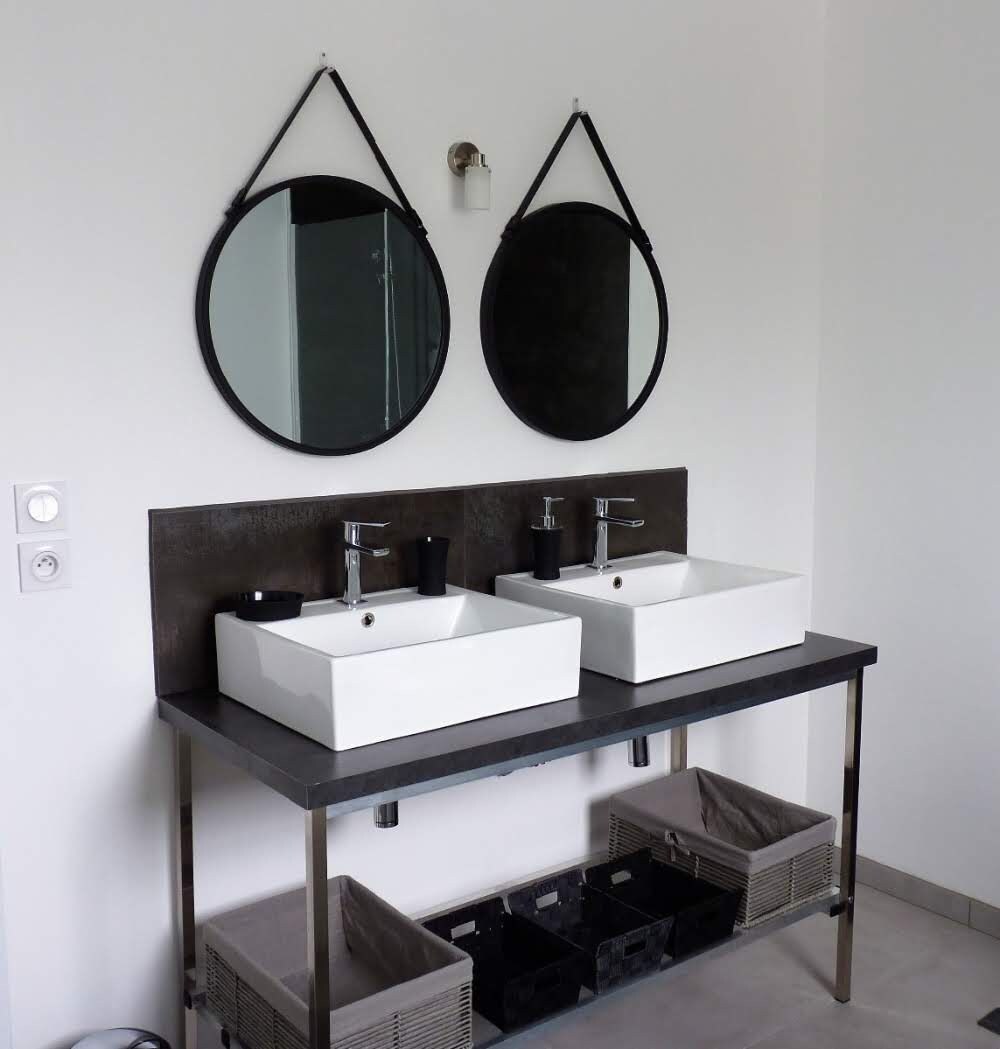 Une salle de bain moderne avec deux vasques blanches, un plan de toilette noir et deux miroirs ronds.