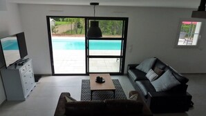A living room with a sofa, a coffee table, a TV, and a sliding glass door leading to a pool. - Air-conditioned villa with heated pool for 9 people (La Motte)