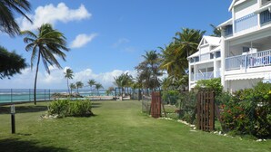 A well-maintained lawn with a fence, palm trees, and a building with balconies. - Very nice F2 facing the sea, direct access to Sainte-Anne beach, swimming pool, tennis (SAINTE ANNE)