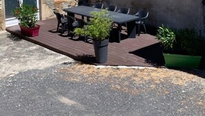 En bygning af sten med tegltag, en trædør og et lille vindue. Der er et bord med stole og plantekasser foran.
