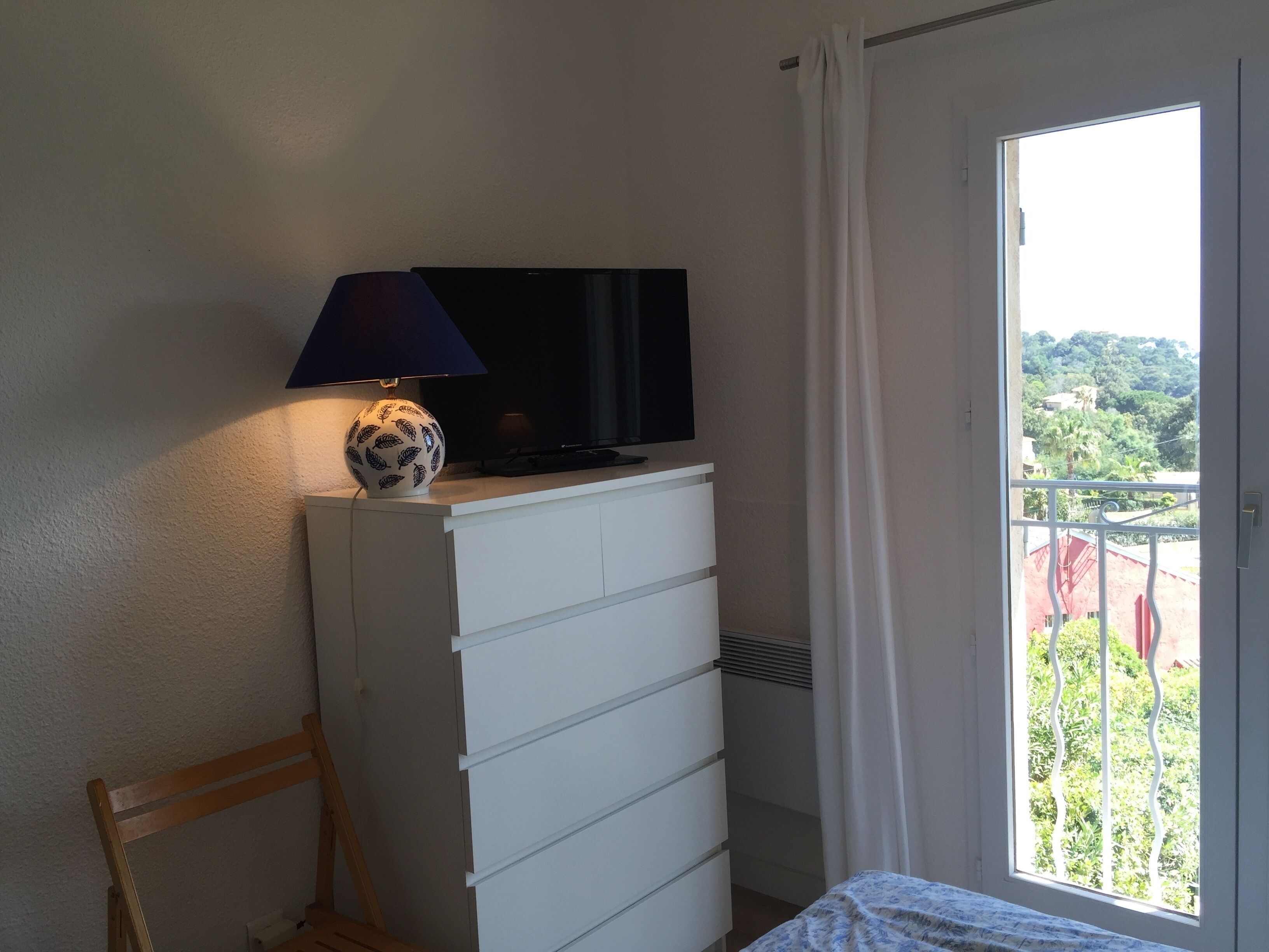 A room with a TV on a white dresser, a lamp, a chair, and a balcony with a view of greenery.