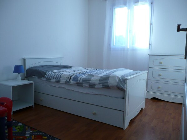 Ein Schlafzimmer mit einem Einzelbett, einem Nachttisch mit Lampe, einem Schminktisch und einem Fenster mit Vorhängen.
