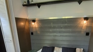 En pænt redt seng med hvide sengetæppe og to puder, et sengebord med en lampe, og en vægmonteret hylde over sengen.