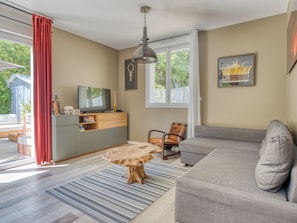 A living room with a grey sofa, a wooden coffee table, a television on a wooden cabinet, and a large window with curtains. - Villa Afrique du Sud - Domaine Le Sherwood (Notre-Dame-De-Monts)
