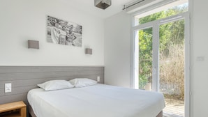 Un dormitorio con cama, mesita de noche, luz empotrada en la pared, ventana con vistas a la vegetación y una fotografía blanco y negro en la pared.