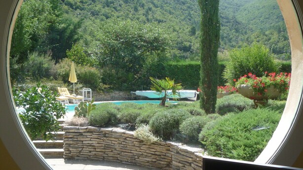 A circular window with a view of a landscaped garden, a swimming pool, and a mountainous backdrop. - Nyons, charming villa in a green setting  (Nyons)
