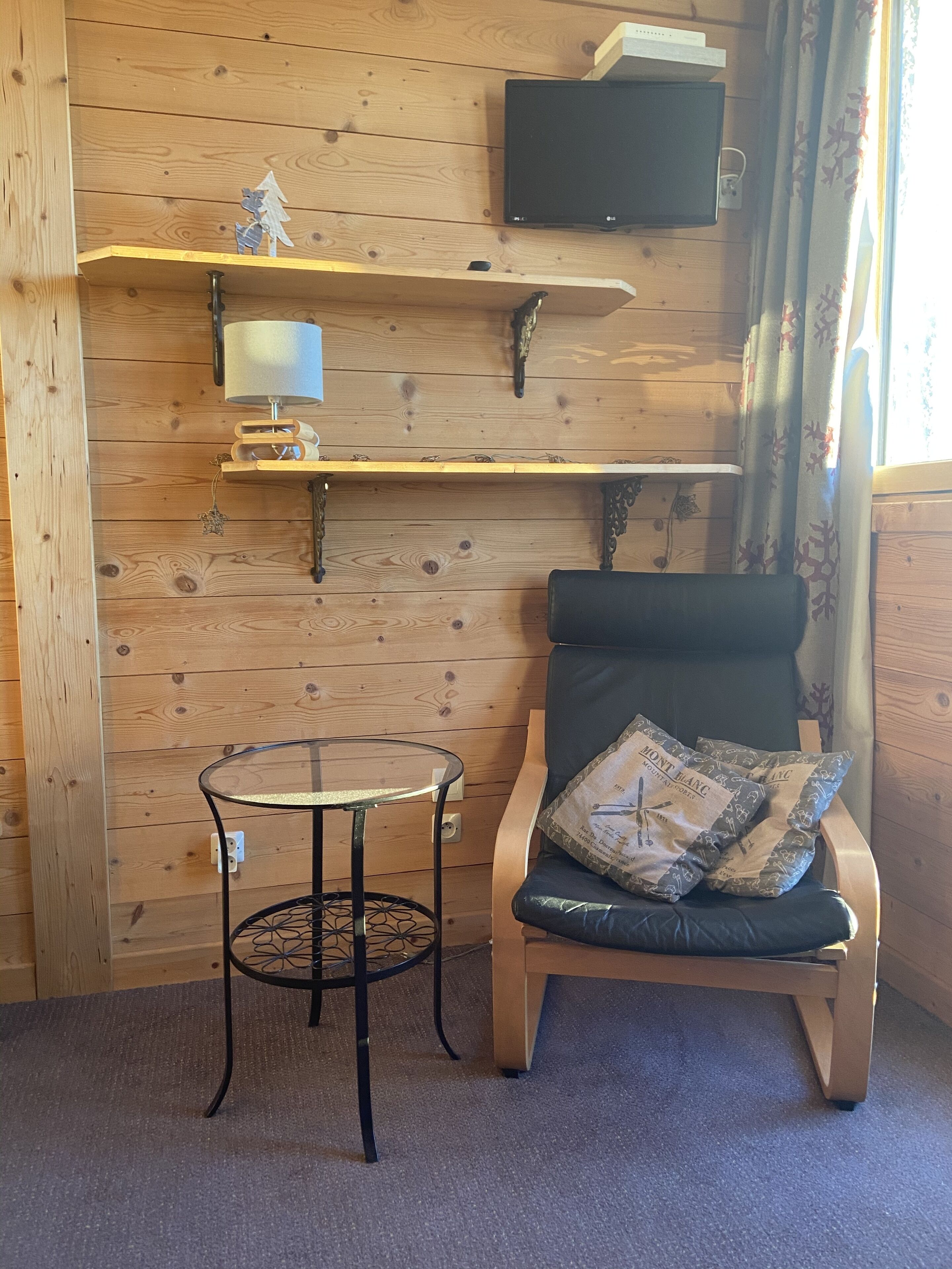 A wooden interior with a TV mounted on the wall, a small table, and a chair with a cushion.