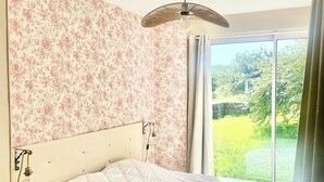 A bedroom with a bed, a headboard, a wall-mounted light fixture, and a view of greenery outside.