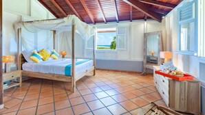 Un dormitorio con cama de dosel de madera, vista a la playa, un armario de madera y suelo de baldosas.