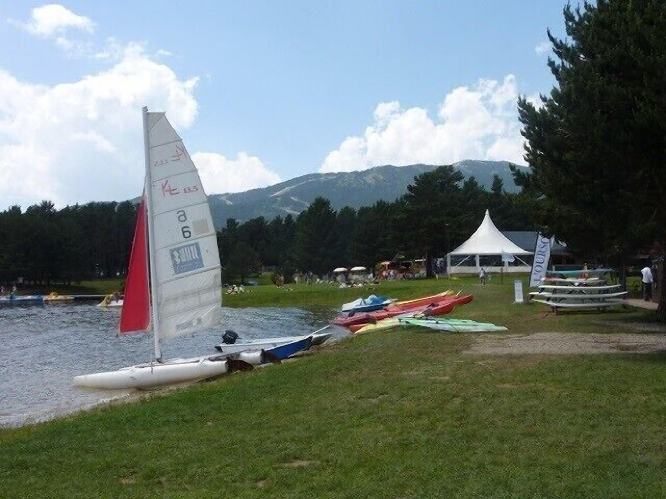 Un paisaje junto al lago con un velero amarrado cerca de la orilla, una tienda de campaña y varias canoas alineadas sobre el césped.