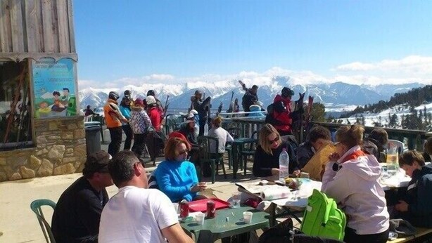 Eine Gruppe von Menschen sitzt an Tischen im Freien, mit einer Berglandschaft im Hintergrund.