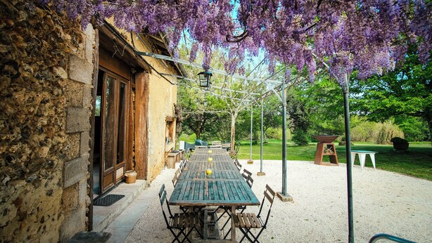 An outdoor dining area with a rustic stone building, a wooden table with chairs, and purple wisteria hanging above. - Exclusive Hamlet and XXL Pool (Vergt)