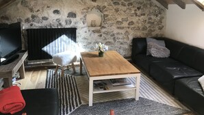 Een knus loft-appartement met een stenen muur, een houten plafond, een zwarte leren bank, een houten salontafel en een televisie op een meubel.
