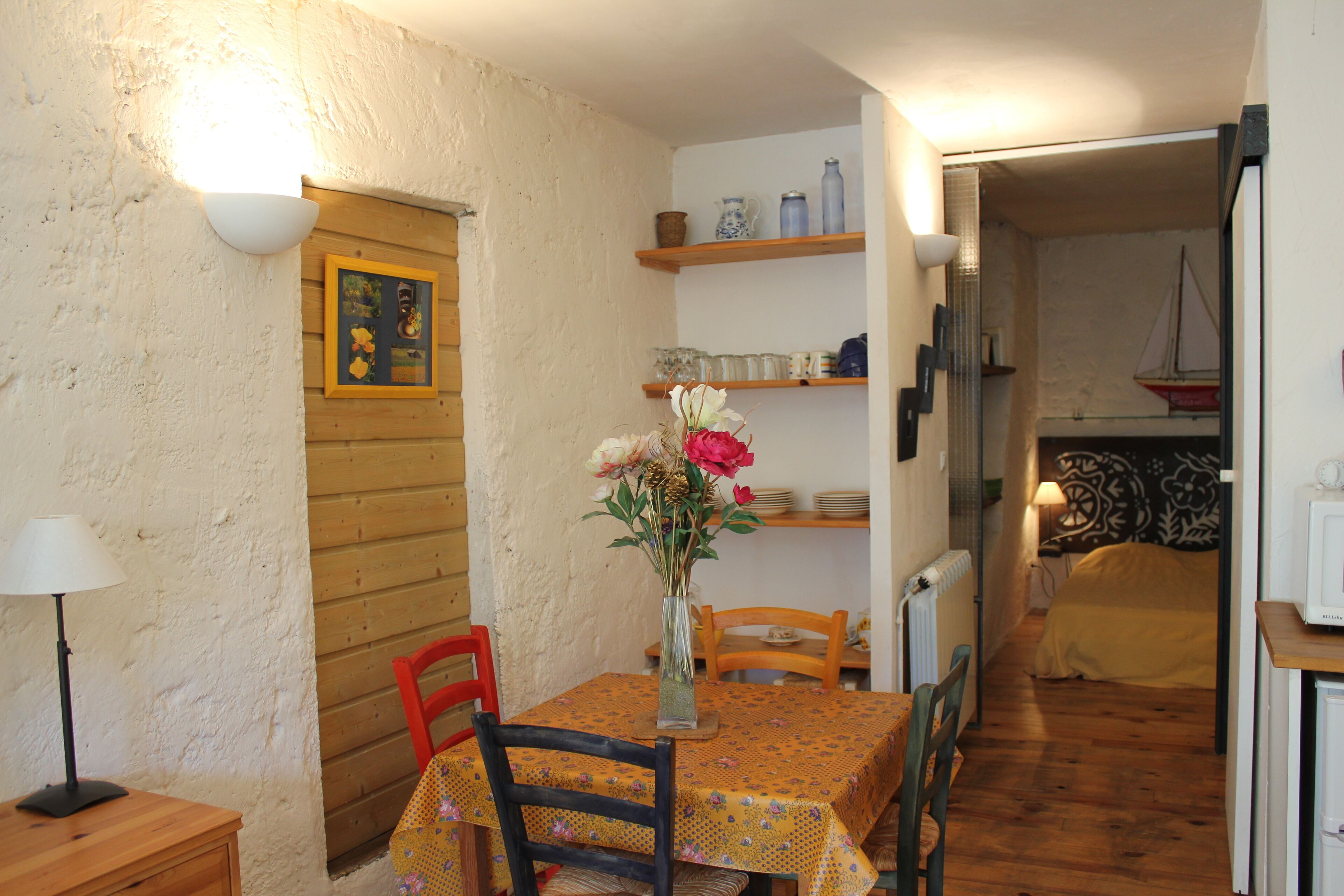 Un coin repas chaleureux avec une table en bois, des chaises et un vase de fleurs. Des étagères au-dessus de la table servent à ranger la vaisselle, et un tableau encadré est accroché au mur.