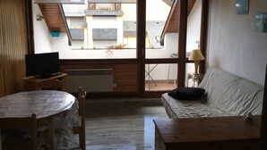 Une pièce avec un téléviseur posé sur une table en bois, un canapé et une fenêtre donnant sur un immeuble.