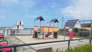 A playground with a slide and climbing structures, surrounded by a fence and a building in the background.