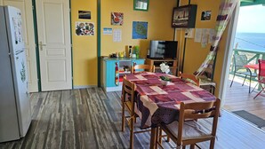 Un'accogliente zona pranzo con un tavolo e sedie in legno, un piccolo mobile con televisore e vista sul mare attraverso una finestra con tende.