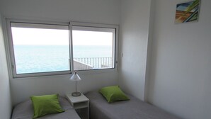 Een kleine kamer met twee éénpersoonsbedden, een nachtkastje, een lamp en een raam met uitzicht op zee.