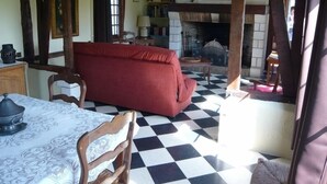 A room with a checkered floor, a red sofa, a wooden table with a patterned tablecloth, and a fireplace with framed pictures above it. - TRADITIONAL GITE NORMAND (La Noë-Poulain)