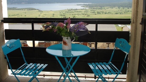 Een balkon met twee blauwe stoelen en een tafel, een vaas met bloemen en uitzicht op een meer en bergen.