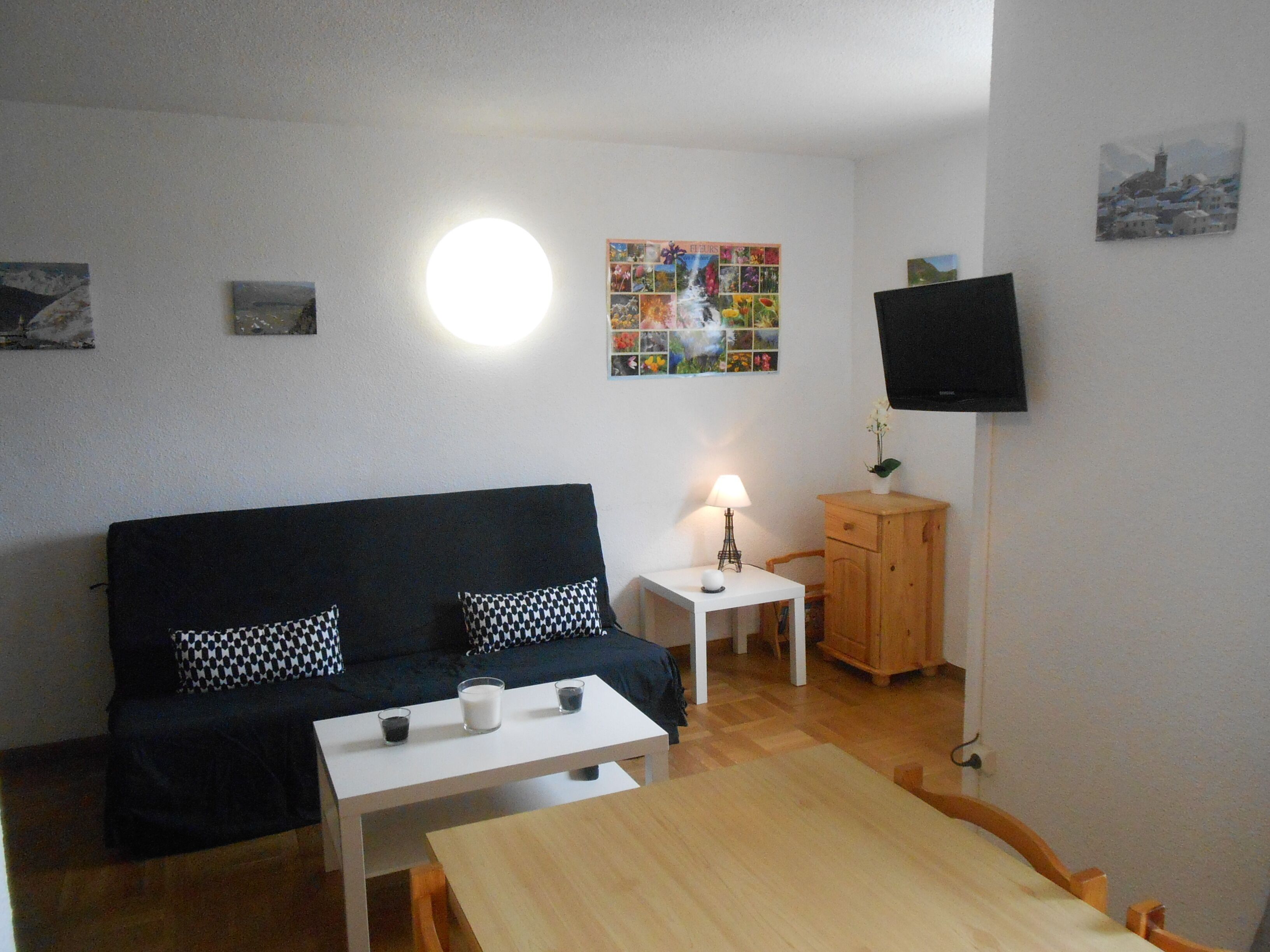A small, furnished room with a black sofa, a white table, a wooden cabinet, a lamp, and a flat-screen TV mounted on the wall.