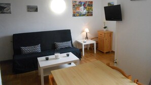 Een kleine, gemeubileerde kamer met een zwarte bank, een witte tafel, een houten kast, een lamp en een flatscreen televisie die aan de muur is bevestigd.