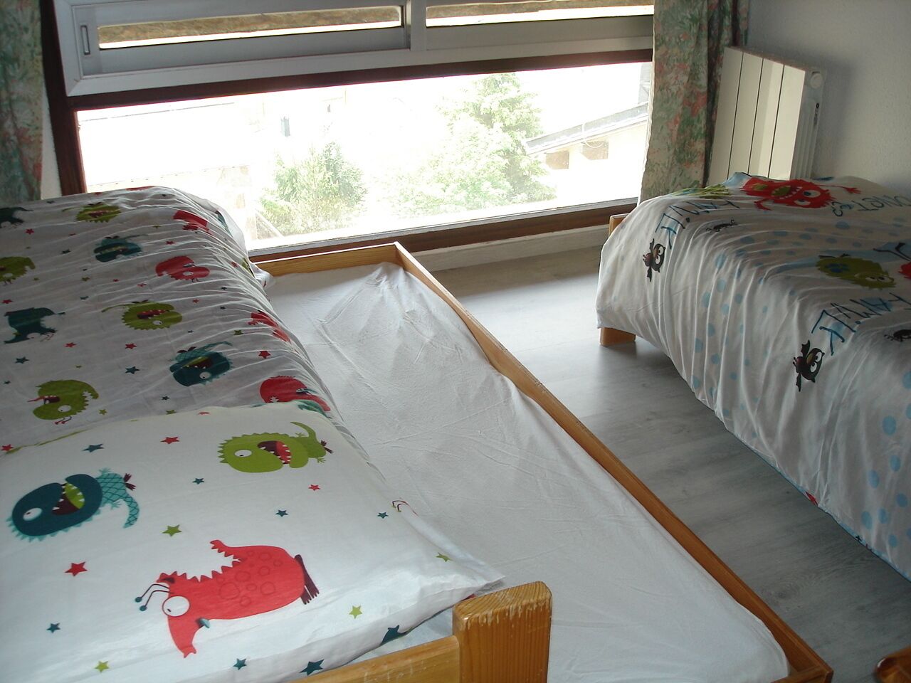 A bedroom with two single beds, each with a colorful bedspread featuring cartoon characters. There is a window with a view of trees and buildings outside.