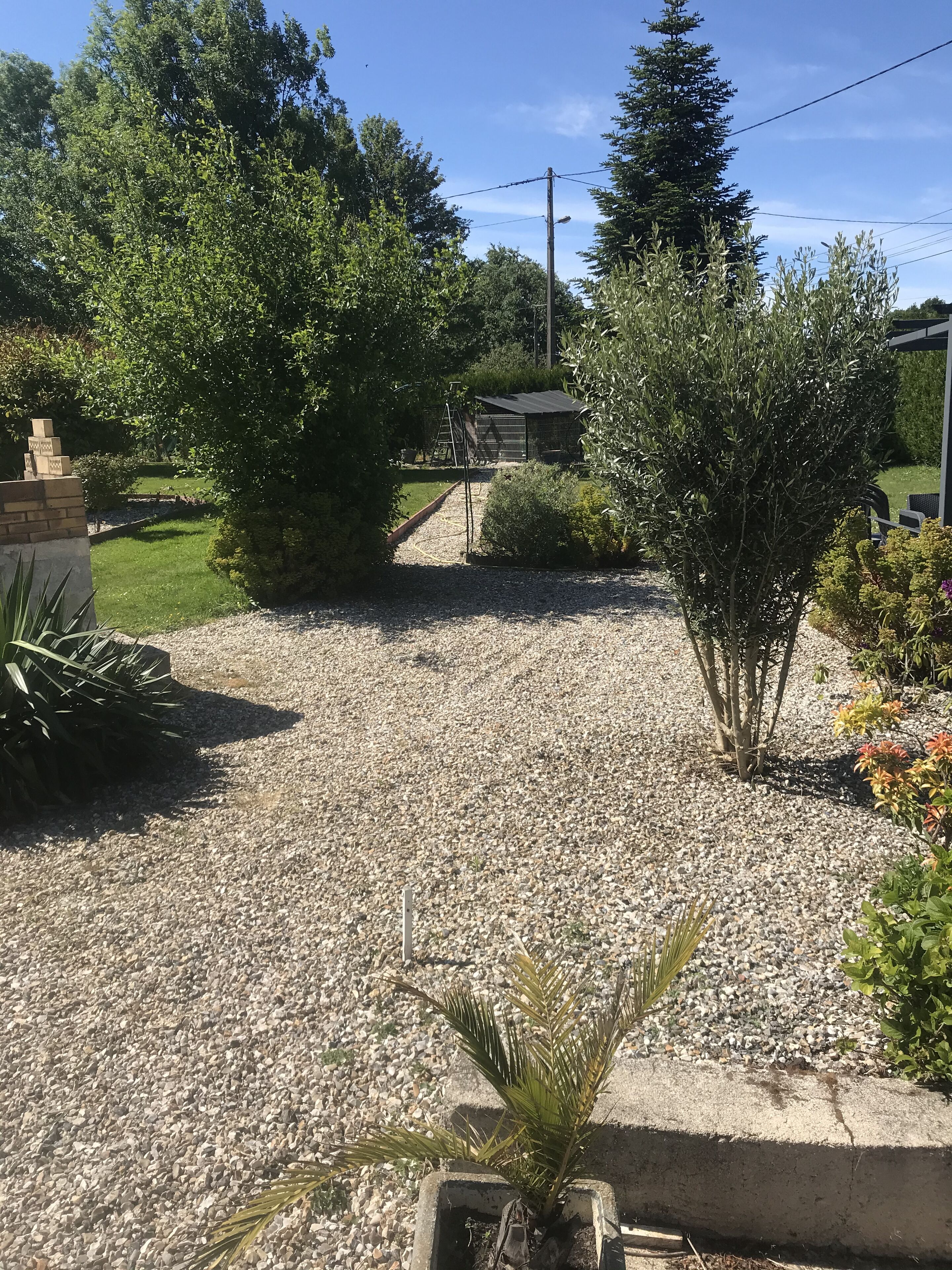 Une allée de gravier mène à une maison dotée d’un petit jardin, d’une clôture en métal et surplombée d’un ciel dégagé.