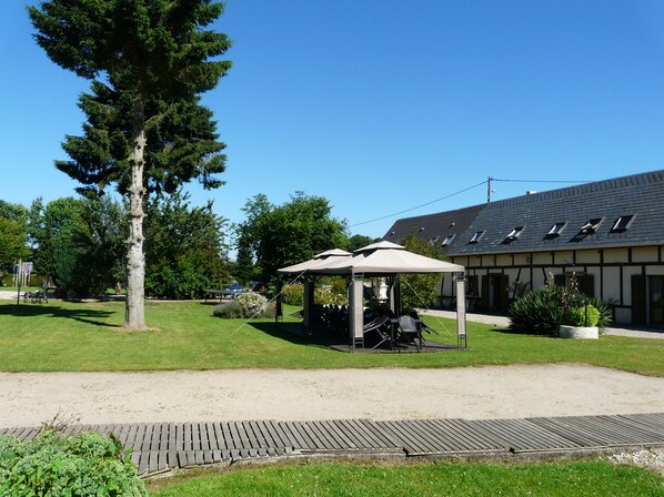 A covered outdoor seating area with a table and chairs, a large tree, and a building with a sloped roof in the background.