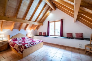 A bedroom with a sloped ceiling, wooden beams, a bed with a patterned bedspread, a window with curtains, and a shelf with decorative pillows. - CAUTERETS Chalet La Grange (sleeps 2–11) in a quiet neighborhood, with parking (Cauterets)
