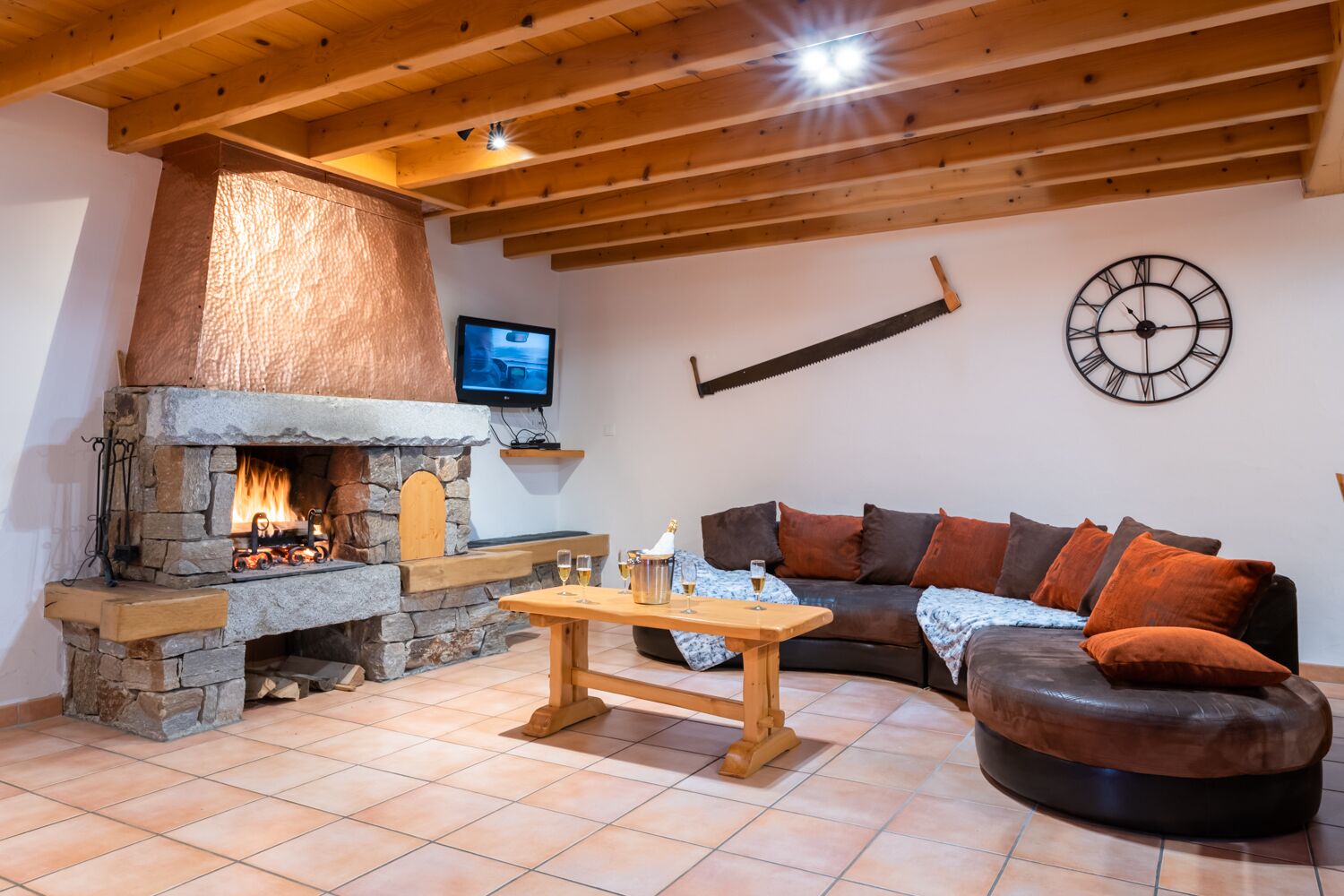 A cozy living room with a stone fireplace, a wooden ceiling, a television mounted on the wall, and a large clock.