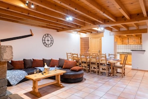 A cozy living room with a wooden ceiling, a stone wall, a sofa with cushions, a wooden table with a bottle and glasses, and a dining area with chairs and a table. - CAUTERETS Chalet La Grange (sleeps 2–11) in a quiet neighborhood, with parking (Cauterets)