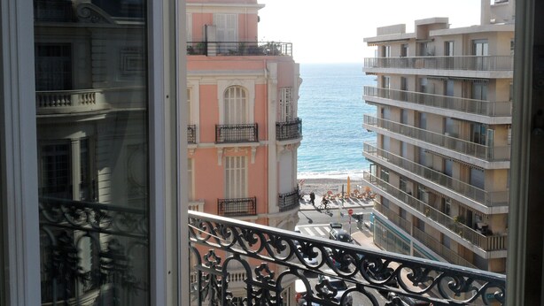 Ein Balkon mit schmiedeeisernen Geländern, der auf eine Küstenstraße mit Gebäuden und Meerblick blickt.