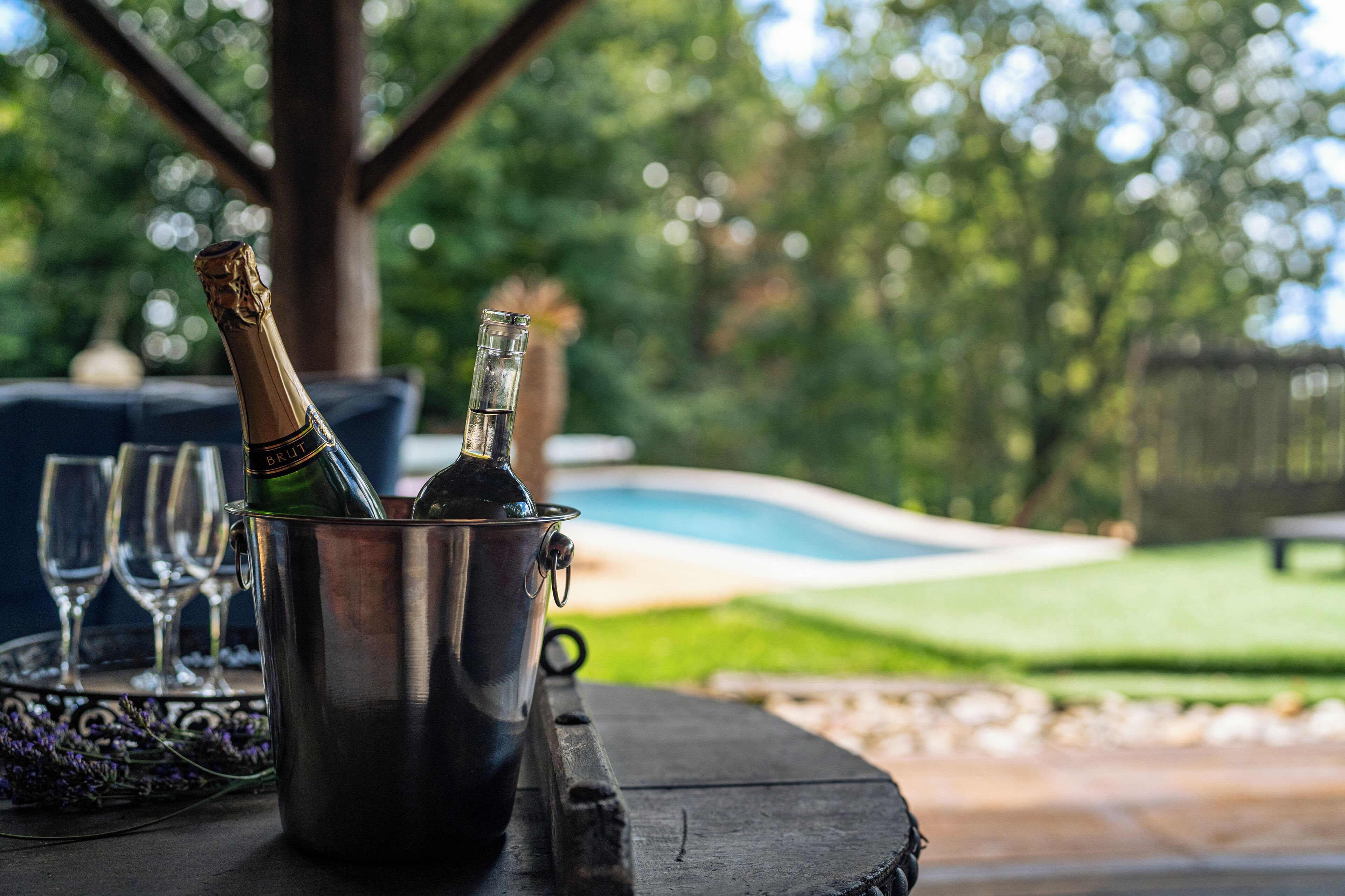 A bottle of champagne in a metal bucket on a wooden table with a pool and lawn in the background.