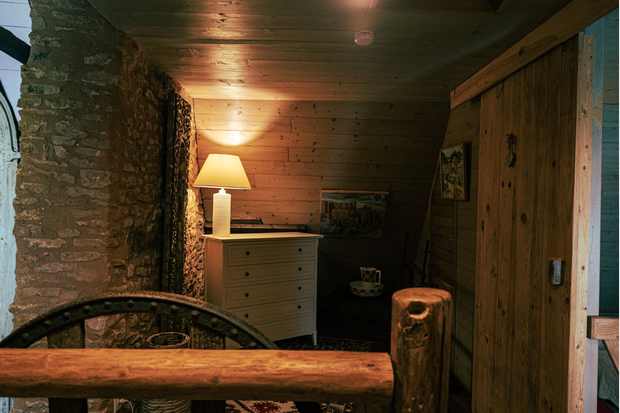 A rustic room with a wooden ceiling, a stone wall, a wooden dresser, a lamp, and a wheel.