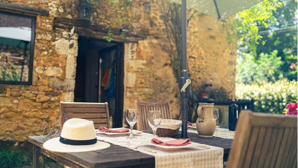 A rustic outdoor dining area with a stone wall, wooden furniture, and a table set with a hat, wine glasses, and a pitcher. - Charm & Tranquility in the heart of Périgord Noir with private heated pool (Le Bugue)
