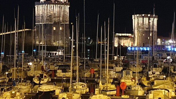 A marina with numerous boats docked, a prominent stone tower, and a smaller tower with illuminated blue lights.