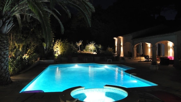 Une piscine illuminée la nuit, entourée de palmiers et bordée d’un bâtiment orné d’arcades.