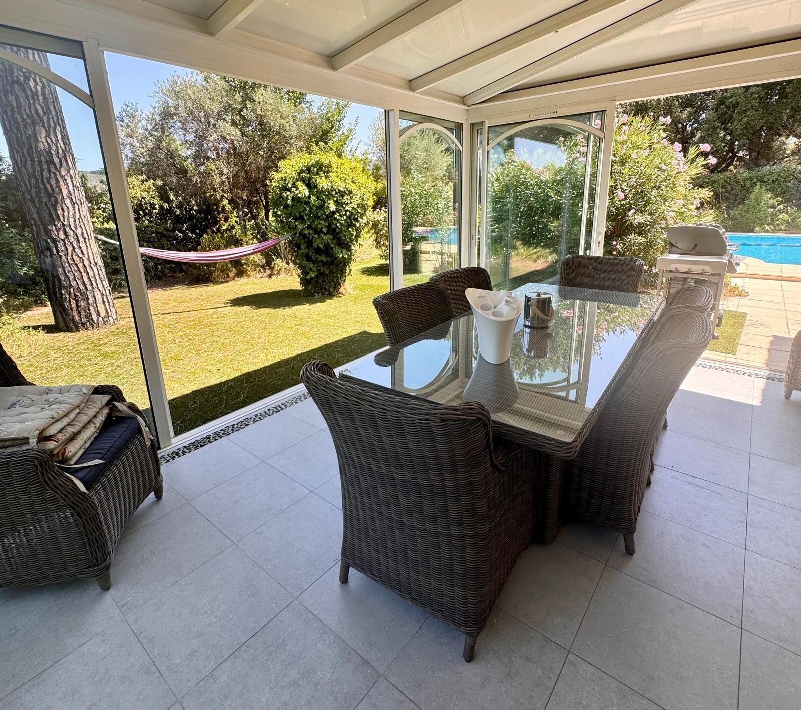 A covered patio with a glass table and wicker chairs, overlooking a pool and garden.