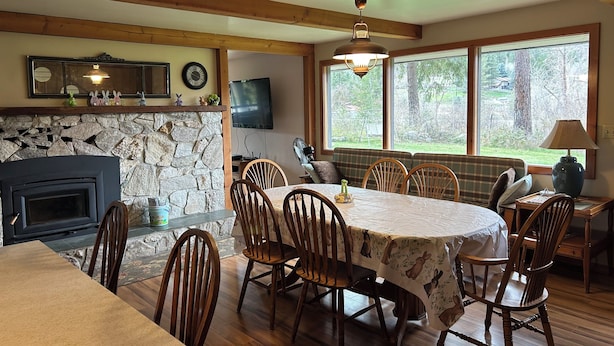 A dining area with a stone fireplace, wooden chairs, a table with a bunny-themed tablecloth, and a view of the outdoors through large windows.